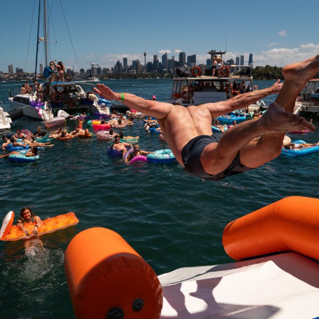 A person jumps off an inflatable slide into a lively body of water filled with people on inflatables, alongside boats and a vibrant city skyline. In the background, a luxury yacht hire Sydney adds to the festive atmosphere.