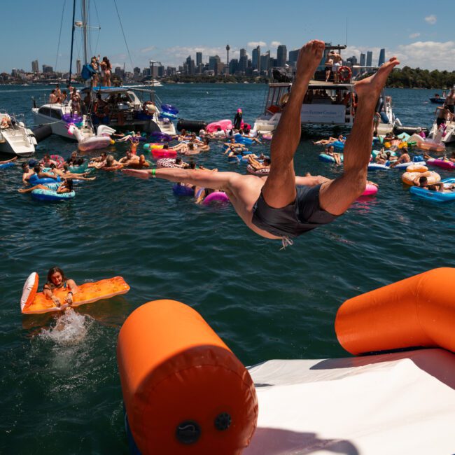 A man is mid-air after jumping off an inflatable structure surrounded by people on floats and boats on a sunny day, with a city skyline in the background. It feels like the perfect setting for a Catamaran party Sydney would host.