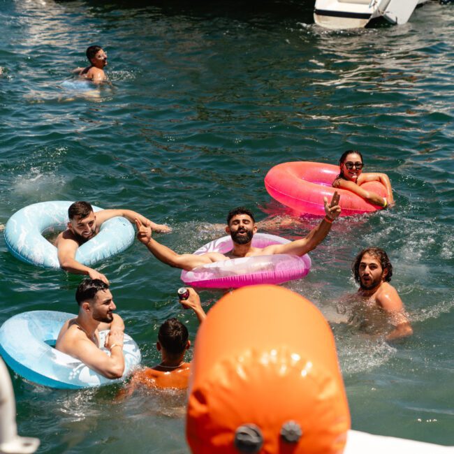 A group of people is enjoying a sunny day in the water, floating on colorful inflatable tubes. They are smiling, waving, and playing in a vibrant and lively setting. The water is clear, with the "Sail Gustavo" and "Capture Studio" logos visible on some of the tubes. A boat is also seen in the background.