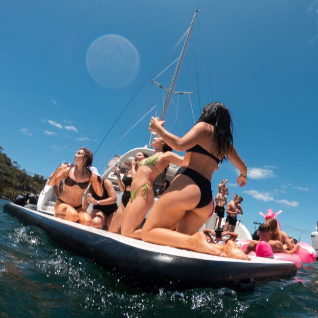 A group of people in vibrant swimwear enjoys themselves on a boat in a sunny, outdoor water setting. Some are dancing, while others are relaxing. The boats are surrounded by clear blue water under a bright sky with a few clouds gently drifting by.