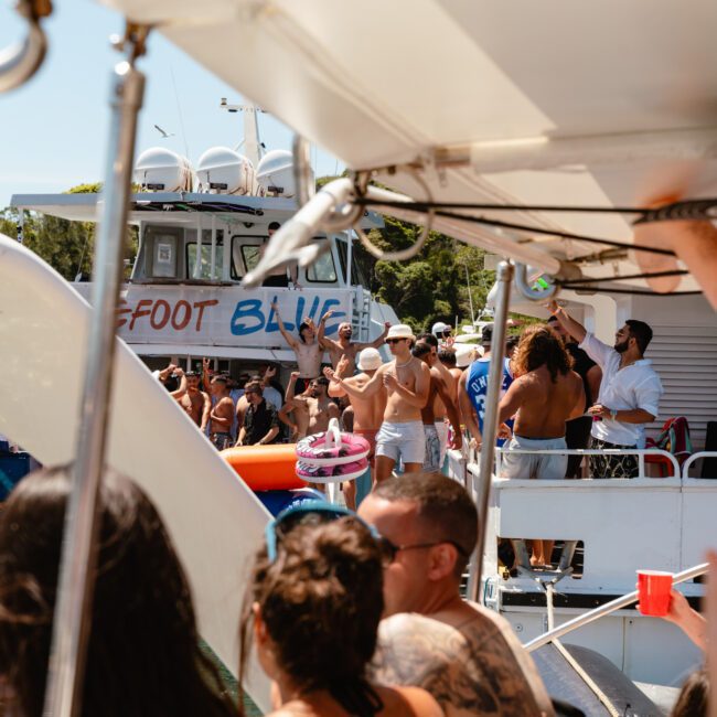 A lively scene on a boat party with people socializing and enjoying drinks. Several individuals are pictured in swimwear on the deck, while a boat named "Bigfoot Blue" floats in the background. The sky is clear, showing a beautiful sunset and the weather appears sunny.