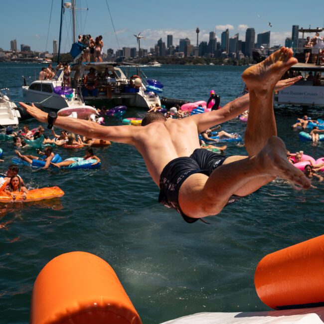 A person jumps off a floating platform into a lively body of water filled with people on inflatables, boats in the background, and a city skyline visible. The scene is buzzing with energy, perfect for those seeking Corporate boat events Sydney or even considering Private yacht charter Sydney Harbour.