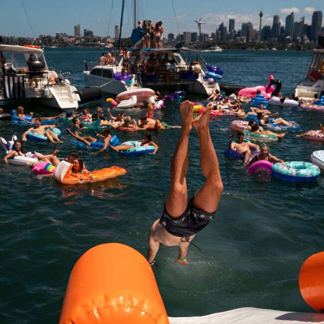 People float on pool inflatables and swim near docked boats in a busy harbor. A person is diving into the water with the skyline in the background, creating a lively atmosphere perfect for corporate boat events Sydney.