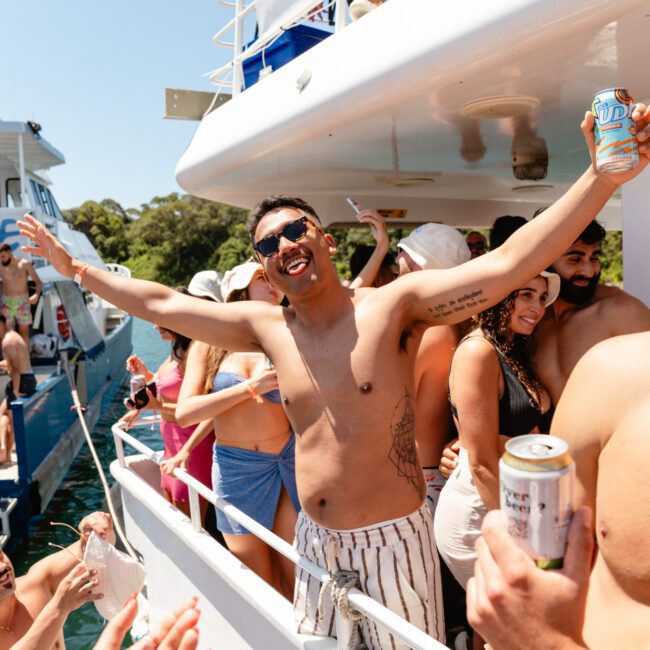 A man wearing sunglasses and striped shorts, with no shirt, spreads his arms wide, smiling, on a crowded boat. He holds a can while others around him also enjoy their drinks. The boat is docked by a sunny coastline with lush greenery in the background.