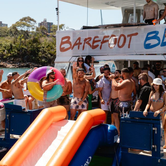 A group of people enjoying a sunny day on a boat named "Barefoot Blue." Some are standing on the deck, waving, while others are in the water or preparing to slide down an inflatable slide into the lake. The background features trees and buildings.