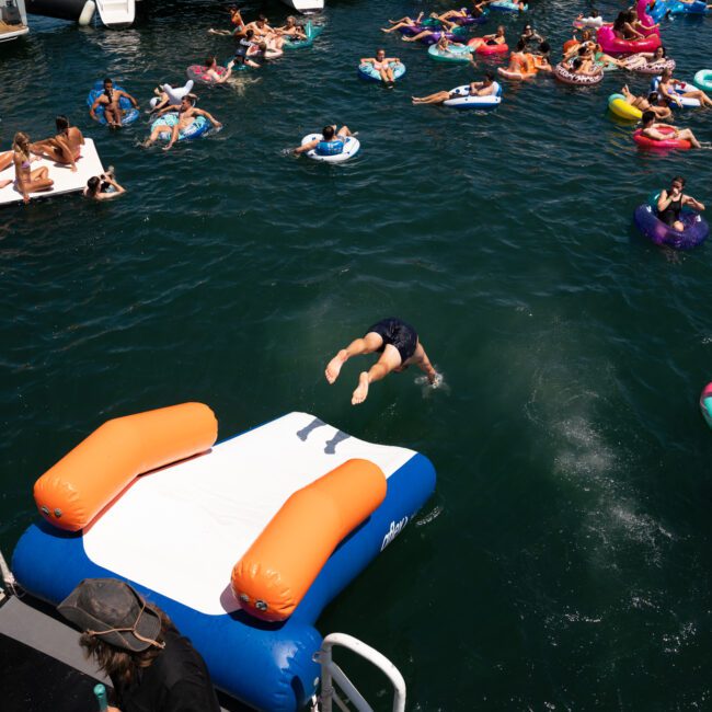 People are enjoying a sunny day on the water, using various inflatables. One person is diving off a large inflatable platform while others float nearby, all under the backdrop of a luxurious yacht hire in Sydney.