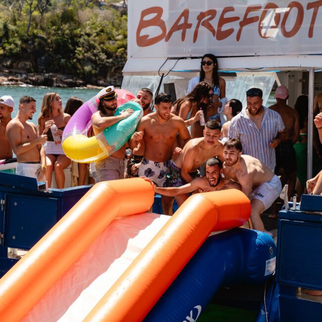 A group of friends gathered on a boat enjoying a lively party. Several are getting ready to go down two inflatable slides into the water. Some wear swimwear and hold colorful pool toys. In the background, there's a lush green landscape and the boat's festive banner flutters in the breeze.