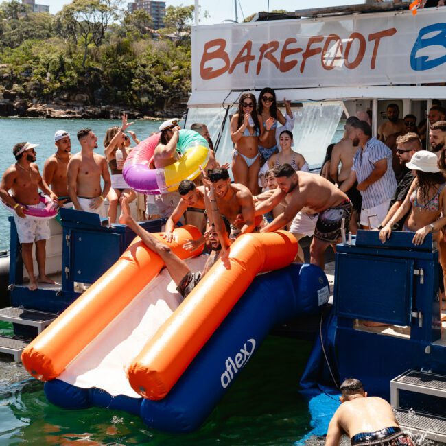 A lively group of people in swimsuits enjoys a boat party on a sunny day. Several individuals are at the back of the boat near an inflatable water slide. One person slides into the water while others cheer and watch, adding to the festive atmosphere. The boat is anchored in a picturesque area.
