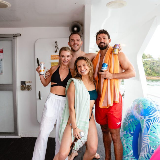 Four people smiling and posing on a boat. Two women, one in shorts and a bikini top, the other in a vibrant bikini with a cover-up, are standing in front. Two men behind them, one in trunks and the other in shorts with a towel over his shoulder. They are holding drinks.