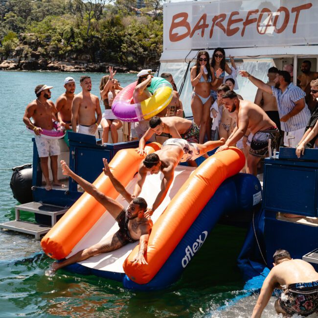 A group of people wearing swimsuits is enjoying a sunny day on a boat named "Barefoot Blue." Some are using an inflatable slide to enter the clear water, while others watch or prepare to slide. The background features serene trees and a rocky shoreline, creating an idyllic summer scene.