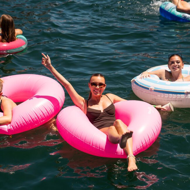 Three people float on inflatable rings in a body of water near a luxury yacht hire Sydney. Two sit on pink rings, and one is on a white ring with blue patterns. They are enjoying the sunny weather with smiles and raised arms.