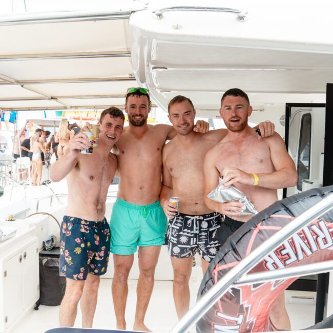 Four shirtless men stand closely together on a boat, each holding drinks and smiling at the camera. Behind them, more people are visible on a nearby boat and in the water. The lively scene suggests a fun, social gathering on a sunny day filled with laughter and camaraderie.