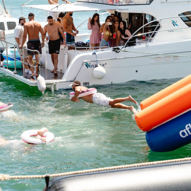 A group of people are enjoying a sunny day on a boat. Some are in the water, with one person diving off from a large inflatable slide, while others watch from the deck. Pool floats add to the fun as the boat is docked in calm, turquoise waters.