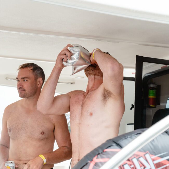 Two shirtless men are on a boat. One man on the left, holding a drink, has a pensive expression. The other man on the right is drinking from a bag and tipping his head back. The man on the right also carries a drink in a koozie at his waistline, enjoying the refreshing breeze.