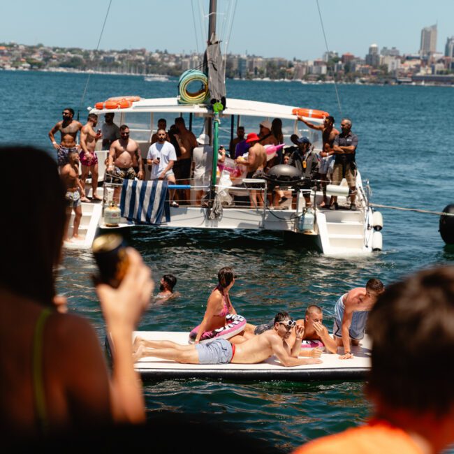 A group of people enjoying a sunny day on a boat and an inflatable float in the water. Some are lying down sunbathing, others are seated or standing, socializing, and holding drinks. The backdrop features a stunning city skyline and blue water.