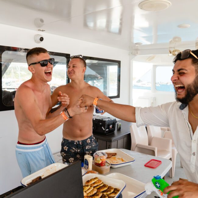 Three men share a laugh on a boat. Two of them, shirtless and wearing sunglasses, stand close together with one patting the other's chest. The third man, in a white shirt and sunglasses, extends his hand towards them while plates of food are laid out on the table, adding to their joyous moment.