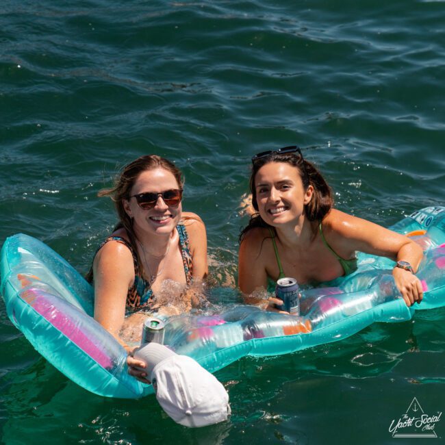 Two women are floating on an inflatable raft in the water, smiling at the camera. Both are holding cans of beverages and wearing sunglasses, enjoying a day of luxury yacht hire Sydney.