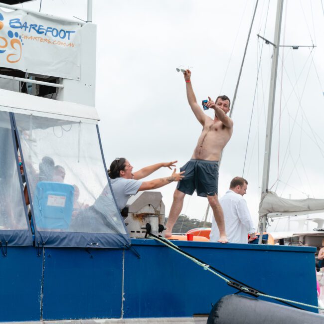 A shirtless man stands on the edge of a blue boat, striking a celebratory pose with one arm raised and holding a phone. Another person, partly obscured, reaches out towards him. A catamaran is in the background, and several enthusiastic individuals are visible on the boat enjoying their time.