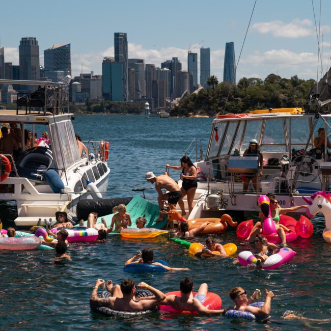 People enjoying a sunny day on inflatable floaties in the water near docked boats, with a vibrant city skyline in the background, as a DJ boat hire Sydney event sets the perfect soundtrack.