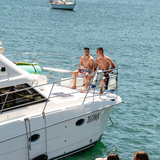 Two men in swim trunks sit on the bow of a white boat named "Big God," enjoying sunny weather. The boat is on calm, blue water with another vessel visible in the background. The image has watermarks reading "SAIL Gustavo" and "Leonardo Brazil.