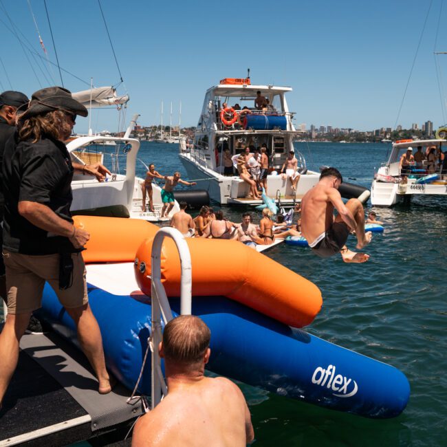 People are enjoying a sunny day on boats and inflatables in the water. Some are jumping in, while others relax onboard during a private yacht charter on Sydney Harbour.