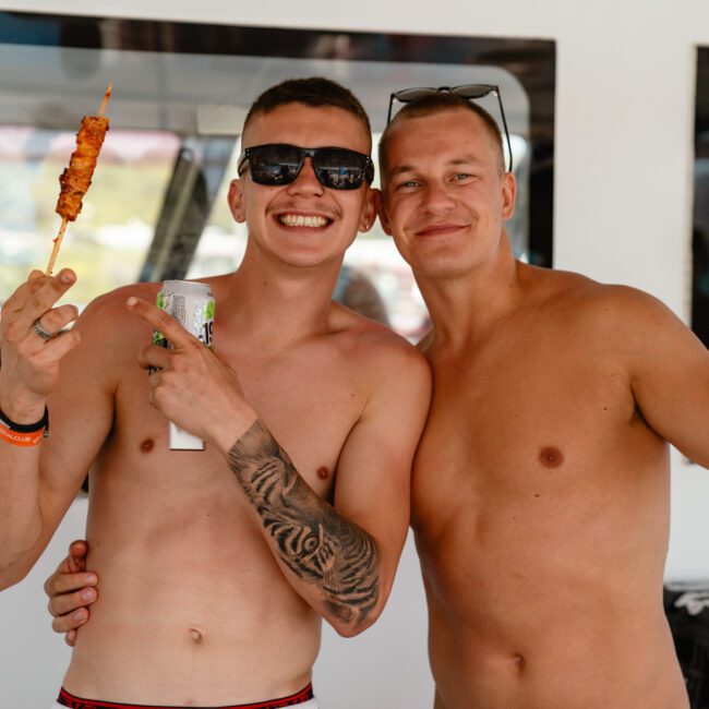 Two shirtless men with tattoos, wearing sunglasses, smile and pose for a photo while holding grilled skewers. They stand in front of a white backdrop with the text "SAIL GUSTAVO" visible in the lower-left corner, capturing a perfect summer vibe.