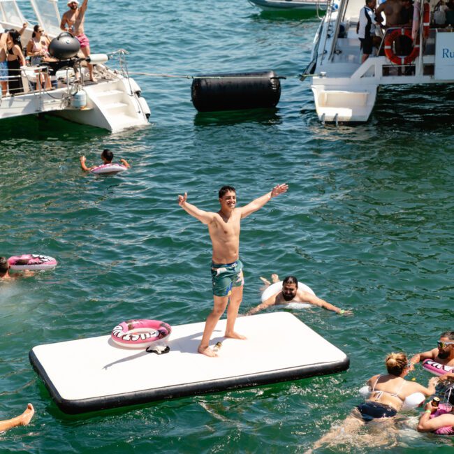 A man stands on a floating platform in the middle of a body of water with his arms raised. Surrounding him are people swimming and socializing. Boats are visible in the background, alongside additional participants enjoying the vibrant aquatic scene.