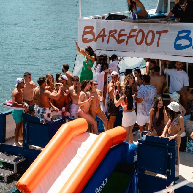 A group of friends enjoying a sunny day on a boat named 'Barefoot Blue.' The boat features a slide extending into the water. Individuals are socializing, dancing to upbeat music, and sitting on the upper deck. The scene is lively and cheerful, with sparkling water in the background.