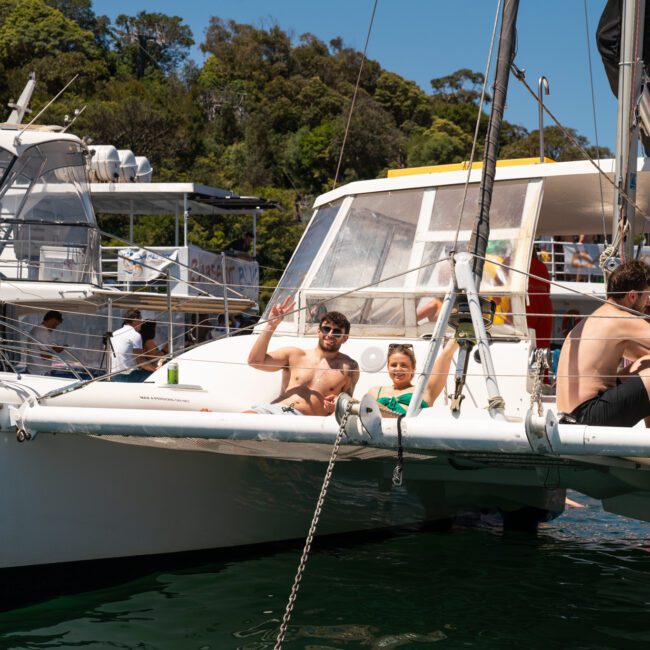 Two people sit at the front of a white catamaran, one waving. Another person is seated near the boat's mast. Other boats and a wooded shoreline are visible in the background under a clear sky, perfect for a Sydney boat party hire.