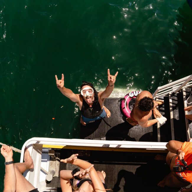 A group of people are on a boat, enjoying a sunny day. One person in the water wearing a snorkel mask is making a rock and roll hand gesture towards the camera. The others are on the boat's ladder and deck, smiling and interacting energetically. The clear green water adds to the tropical vibe.