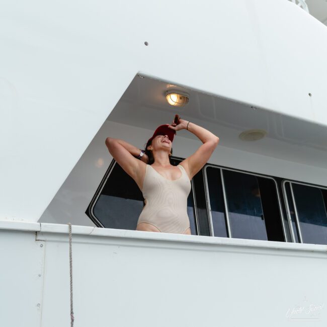 A woman in a beige swimsuit and red visor stands with her hands behind her head on a luxurious yacht, looking up. The yacht's white structure elegantly frames her, contrasting beautifully against the clear blue sky.