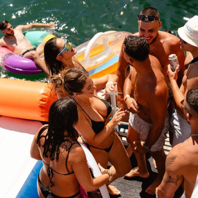 A group of people enjoy a sunny day on a boat party. Some are standing on the deck, chatting and holding drinks, while others relax on inflatable floats in the water. Dressed in vibrant swimwear, they dance joyfully to the upbeat music, adding to the lively and cheerful atmosphere.