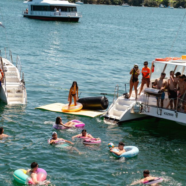 People are enjoying a sunny day on the water near two large boats. Some are floating on inflatable rings, while others are standing on a floating dock between the boats. The water is calm and a third boat with vibrant decorations is seen in the background, adding to the festive atmosphere.