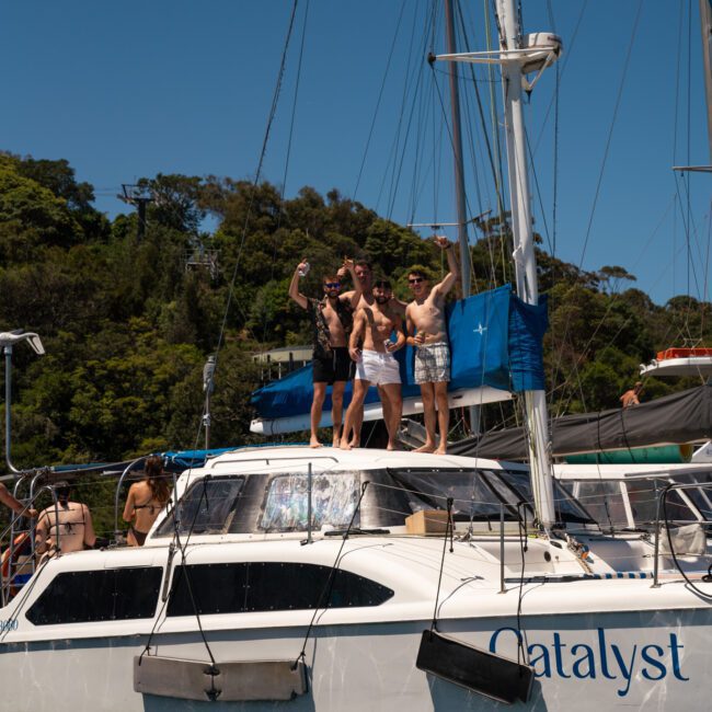 Three people are standing on a yacht named "Catalyst," raising their arms and posing for the camera, enjoying a beautiful catamaran party in Sydney. The boat is on the water with trees visible in the background.