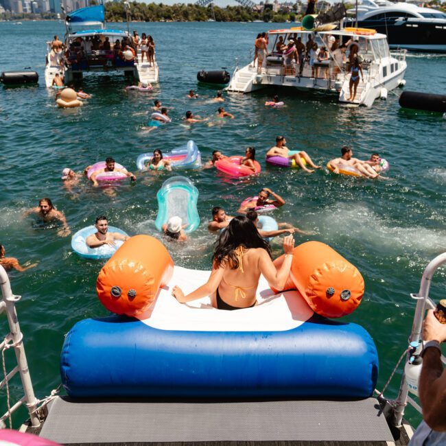 A group of friends is enjoying a sunny day on the water. Some are on boats, while others float on colorful inflatables. A woman is about to slide from a large inflatable slide into the lake. The scene is lively with numerous people swimming and relaxing in the refreshing water.