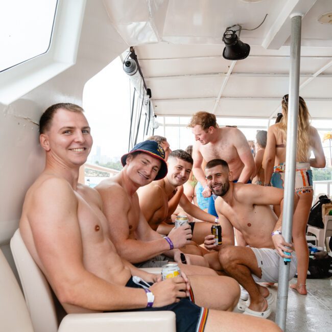 A group of shirtless men sit and smile at the camera on a boat, dressed in swim trunks, some holding drinks. More people are seen standing in the background, enhancing the lively atmosphere. The setting appears to be a fun social gathering or party on the water.