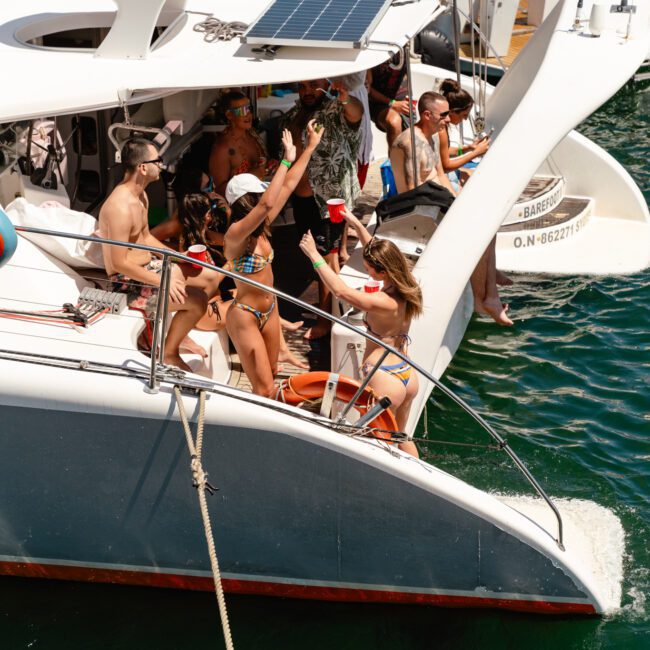 A group of people enjoys a sunny day on a catamaran yacht, wearing swimwear and engaging in various activities like dancing and chatting. Some hold drinks, while others sit with their feet dangling over the water. Laughter echoes as the breeze carries the pleasant scent of the sea around them.