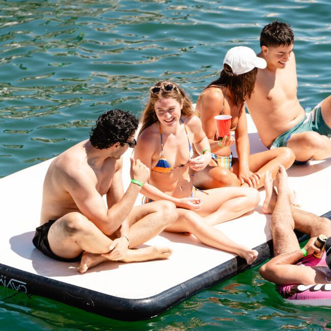 Four young adults in swimwear relax and chat on a floating dock on a sunny day. They are enjoying themselves, with smiles and laughter, against a backdrop of clear, green water. Two logos, "Sal Gustavo" and "Yacht Social Club," are visible in the image.