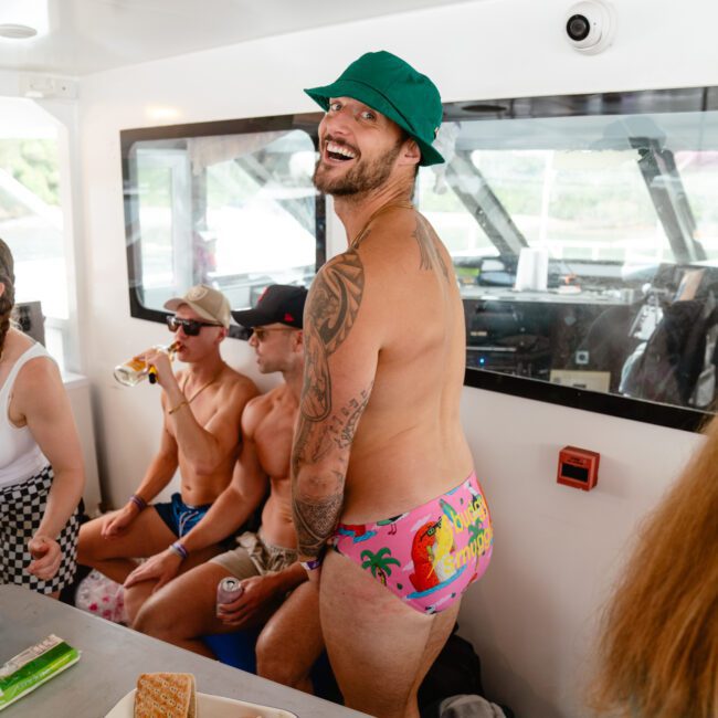 A group of men on a boat enjoy a casual gathering. The focus is on a man standing and smiling at the camera; he is wearing a green hat and colorful swim trunks. Others are seated, some with beverages in hand. The setting is cheerful, relaxed, and picturesque under the sunny sky.