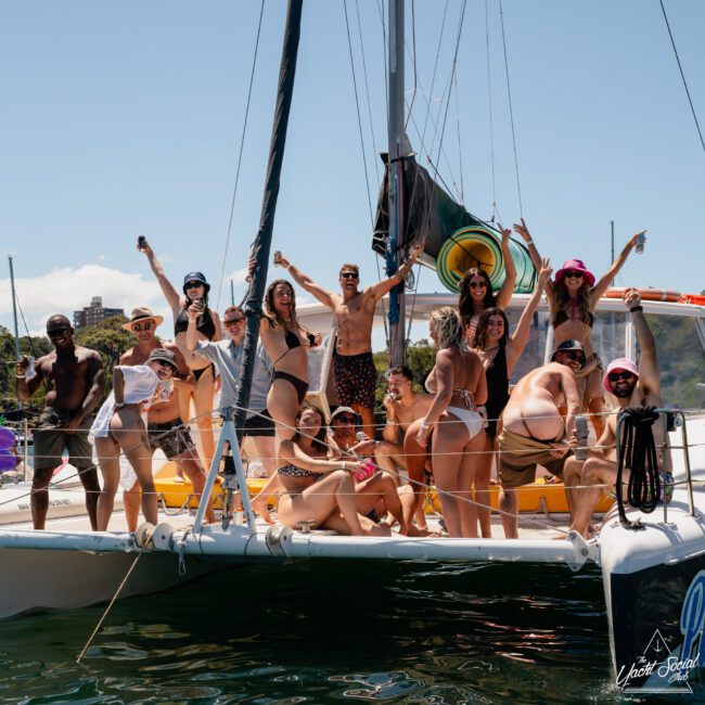 Group of 16 people in swimwear enjoying a day on a catamaran, some holding drinks and posing joyfully. Clear sky and water in the background, perfect for a Catamaran party Sydney.