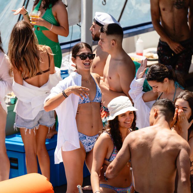 A group of people in swimsuits enjoy themselves on a boat. Some are dancing, others are laughing and socializing under the bright sun. The atmosphere is lively and festive, with the sea and part of the boat visible in the background. A logo in the corner reads "Sail Gustavo.