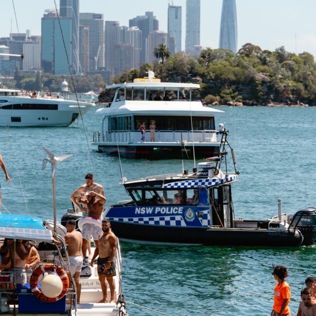 A group of people enjoy a sunny day on boats in a harbor with city skyscrapers in the background. A police boat marked "NSW POLICE" is nearby, and some individuals are interacting with it. The water is calm, reflecting the tall buildings, and there are several trees on a small island.
