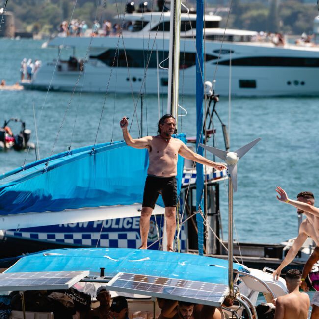 A man stands on the bow of a sailboat, holding onto a railing, wearing swim trunks. The sailboat is filled with people enjoying the sunny day. In the background, other boats dot the water and a city skyline rises majestically. The scene appears lively and celebratory.