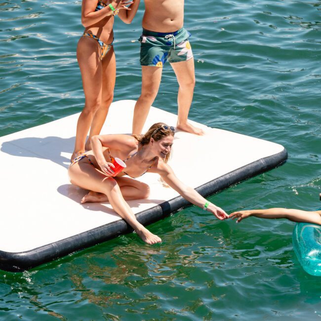 A group of people enjoying a sunny day on the water. Two individuals stand on an inflatable platform with a woman sitting and holding a red cup, reaching out to another woman lounging on a floating tube. They are surrounded by clear, greenish-blue water and laughter fills the air.
