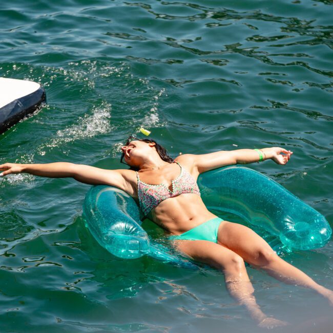 A woman in a colorful bikini is lying on a blue inflatable pool float, joyfully stretching her arms to the sides. The bright, crystal-clear water is splashing nearby, with part of a boat and vibrant palm trees visible in the background.