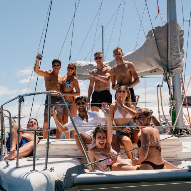 A group of people in swimwear are posing and smiling on a yacht deck under a clear sky, enjoying a lively catamaran party in Sydney.