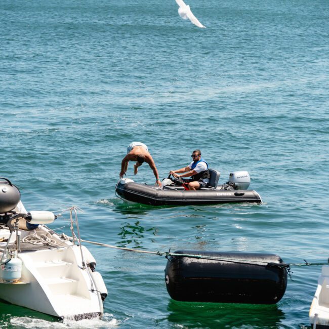 A group of people are enjoying themselves on a catamaran and a motor yacht in a sunny, open-water setting. In the center, a person in swimwear transitions from the yacht to an inflatable boat, assisted by another person seated in the inflatable. Some seagulls are flying overhead at The Yacht Social Club Event Boat Charters.