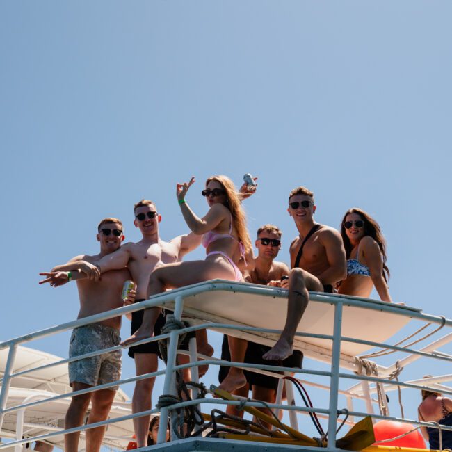 A group of six people poses on the upper deck of a luxury yacht hire in Sydney under a clear blue sky. Some are holding drinks, and one person is making a peace sign.