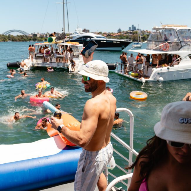 A lively scene on a sunny day with many people enjoying a boat party on the water. Individuals are on paddleboards and swimming around, with boats moored nearby. People are socializing, dancing, and having fun in their swimsuits, with the city skyline visible in the background.