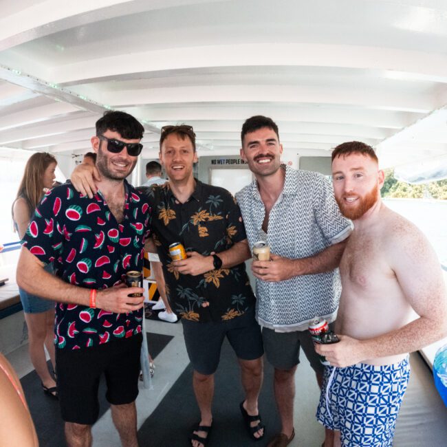 A group of four men smiling and posing for a photo on a boat, dressed in casual summer attire and holding drinks. Two sport sunglasses. The background showcases inflatable pool toys and water, clearly indicating they are enjoying a fun day on the lake.
